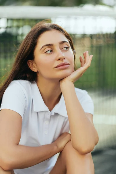 Tennis court in Miami, portrait of dreamy female tennis player with brunette hair wearing white polo shirt and looking away after training, tennis net on blurred background, Florida — Stock Photo