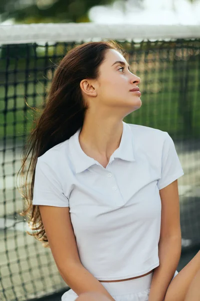 Tennisplatz in Miami, Porträt einer abgelenkten Tennisspielerin mit brünetten Haaren, die weißes Poloshirt trägt und nach dem Training wegschaut, Tennisnetz auf verschwommenem Hintergrund, Florida — Stockfoto