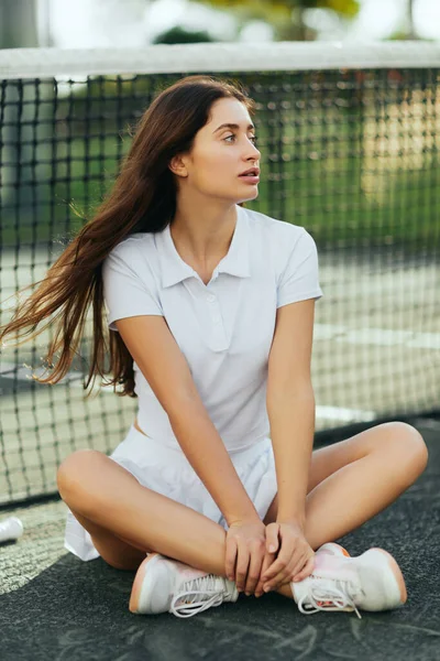 Distraído jugador femenino en la cancha de tenis, mujer joven con el pelo largo sentado con las piernas cruzadas en traje blanco y zapatillas de deporte y mirando lejos cerca de la red de tenis, fondo borroso, Miami, tiempo de inactividad - foto de stock
