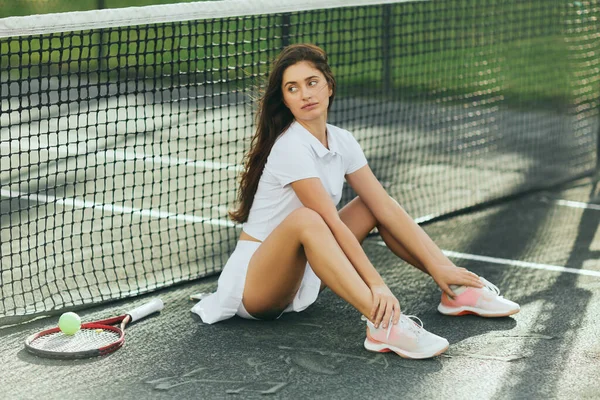 Jugadora de tenis mujer calentando antes del partido, mujer joven con el pelo largo sentado en traje blanco cerca de raqueta con pelota y red de tenis, fondo borroso, Miami, ciudad icónica, pista de tenis - foto de stock