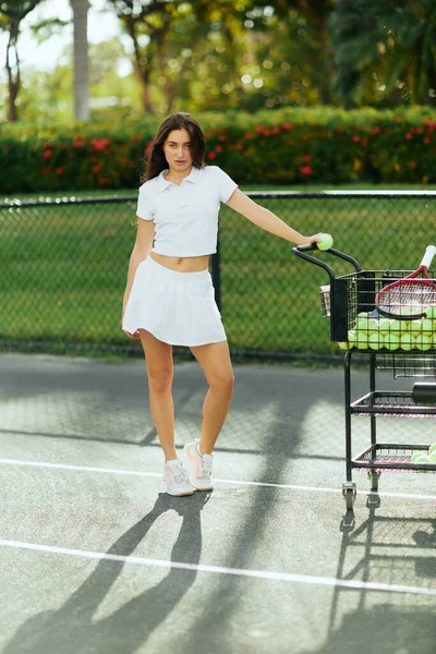 Graziosa tennista, giovane donna sportiva con capelli castani in piedi in abito bianco con gonna e polo vicino al carrello con palle, sfondo sfocato, baciato dal sole, campo da tennis a Miami — Foto stock