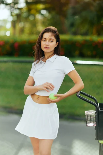 Physical activity, young woman with brunette hair standing in stylish outfit with skirt and white polo shirt near cart and holding ball, blurred background, sun-kissed, tennis court in Miami — Stock Photo