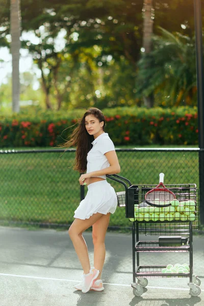 Atlético, sexy jovem mulher com cabelo morena em pé na roupa elegante com saia e camisa de pólo branco perto de carrinho com bolas, fundo borrado, beijado pelo sol, quadra de tênis em Miami — Fotografia de Stock