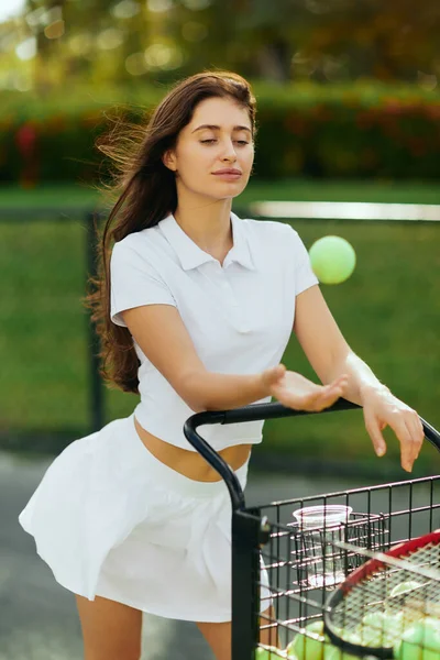 Habitudes saines, jolie jeune femme aux longs cheveux bruns debout en tenue blanche près du chariot et jetant la balle dans l'air, fond flou, court de tennis à Miami, énergie et plaisir — Photo de stock