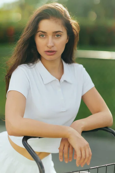 Jolie joueuse de tennis, jeune femme sportive aux cheveux bruns debout en polo blanc près d'une voiturette de tennis, fond vert flou, regardant caméra, court de tennis à Miami — Photo de stock