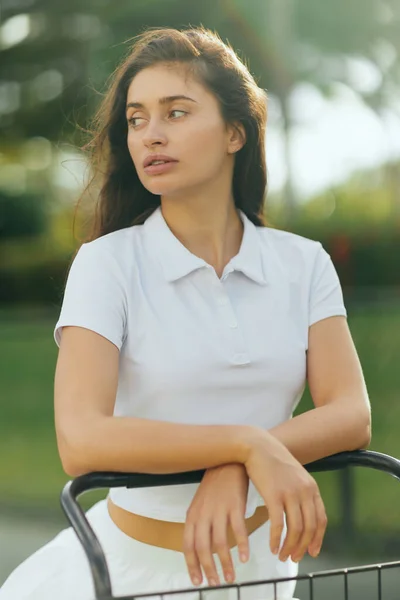 Jogadora de tênis, jovem esportiva com cabelo morena de pé em camisa de pólo branco perto de carrinho de tênis, fundo verde borrado, olhando para longe, quadra de tênis em Miami, cidade icônica — Fotografia de Stock