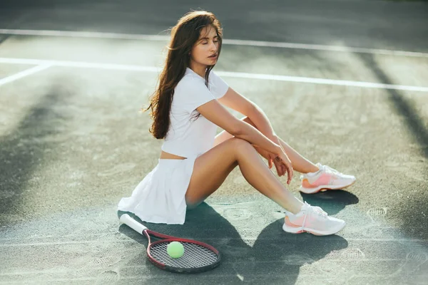Jogadora feminina descansando após o jogo, jovem morena com olhos fechados sentada em roupa branca perto de raquete com bola no asfalto, Miami, quadra de tênis, tempo de inatividade, sombras, dia ensolarado — Fotografia de Stock