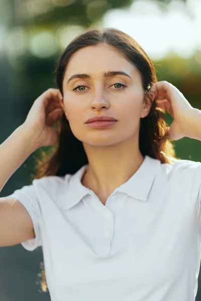 Portrait de jeune femme élégante aux cheveux longs bruns debout en polo blanc et regardant la caméra, fond flou, Miami, Floride, ville emblématique, maquillage naturel — Photo de stock