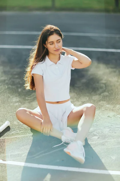 Pensive young woman with long hair sitting in white outfit near tennis racket on asphalt, blurred background, Miami, tennis court, downtime, shadows, sunny day, high angle view, looking away — Stock Photo