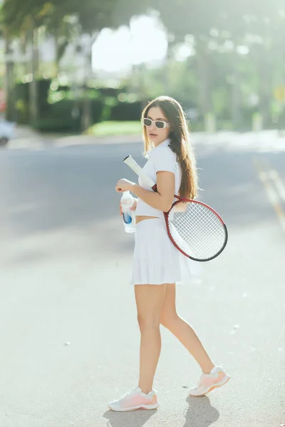 Pretty tennis player, young woman with long brunette hair standing in white sporty outfit and sunglasses with racket and bottle of water on urban street in Miami, blurred background, healthy habits — Stock Photo