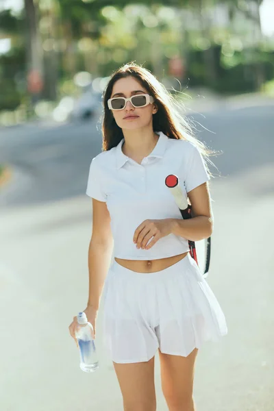 Jugadora de tenis, mujer joven con el pelo largo morena caminando en traje deportivo blanco mientras sostiene raqueta y botella con agua en la calle urbana en Miami, fondo borroso, hábitos saludables - foto de stock