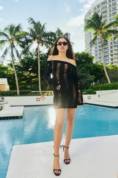 Sexy brunette woman in black knitted dress and sunglasses posing against palm trees and modern hotel building in Miami, vacation, outdoor swimming pool with shimmering water in luxury resort — Stock Photo