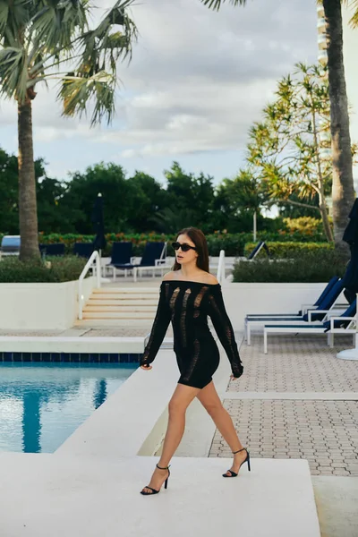 Séduisante femme en robe tricotée noire et lunettes de soleil marchant en talons hauts à côté de la piscine extérieure avec de l'eau chatoyante contre les palmiers à Miami, vacances dans un complexe de luxe, chaises longues — Photo de stock