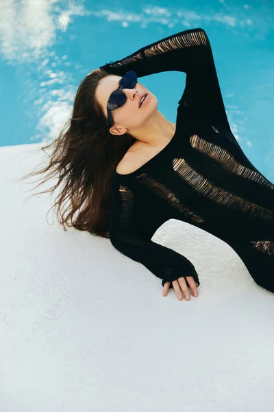 Resort de luxe, femme brune sexy avec peau bronzée en robe tricotée noire et lunettes de soleil posant à côté de la piscine extérieure avec eau chatoyante à Miami, escapade estivale, jeunesse — Photo de stock