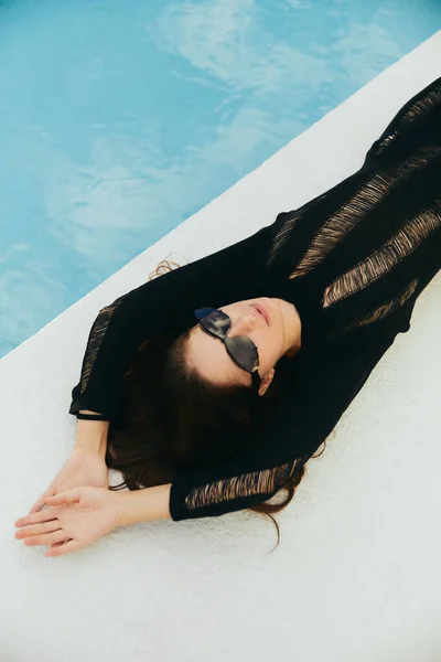 Tentação, resort de luxo, mulher sexy com pele bronzeada em vestido de malha preto e óculos de sol ao lado da piscina exterior com água azul em Miami, vista superior — Fotografia de Stock