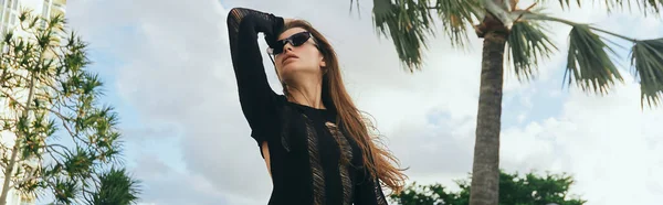 Luxury resort, sexy brunette woman with tanned skin in black knitted dress and sunglasses standing against palm trees and blue sky in Miami, summer getaway, banner — Stock Photo