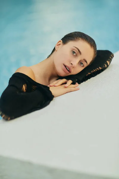 Розкішний курорт в Маямі, красива жінка з засмаглою шкірою в чорному купальнику плаває в громадському басейні, виставляється поруч з басейном і насолоджується літньою відпусткою, без макіяжу — стокове фото