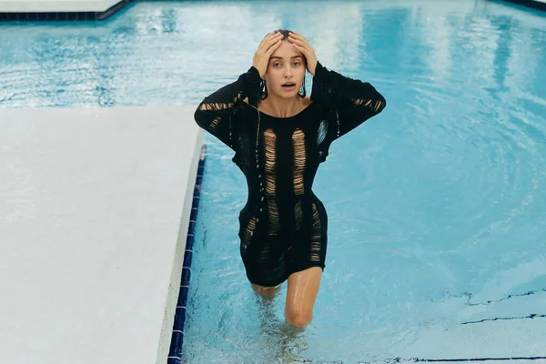 Resort de lujo en Miami, mujer sexy con piel bronceada y vestido negro mojado mirando a la cámara, dentro de la piscina pública, posando y disfrutando de sus vacaciones de verano, sin maquillaje — Stock Photo