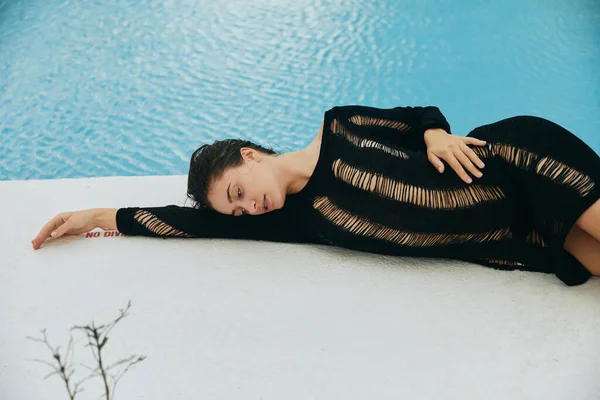 Роскошный курорт, сексуальная женщина в черном вязаном платье, лежащая рядом с открытым бассейном с мерцающей водой в Майами, летний отдых, молодость, отдых у бассейна, загар — стоковое фото