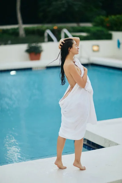 Young sexy woman with wet hair wrapped in white towel standing next to outdoor swimming pool with shimmering water in Miami, summer getaway, youth, poolside relaxation, vacation mode — Stock Photo