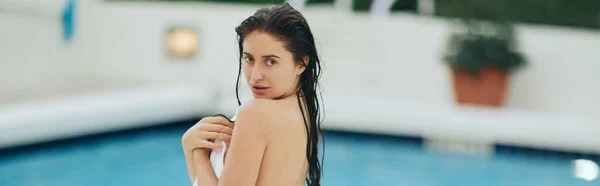 Young brunette woman with wet hair wrapped in white towel standing next to outdoor swimming pool in Miami, summer getaway, youth, poolside relaxation, vacation mode, looking at camera, banner — Stock Photo