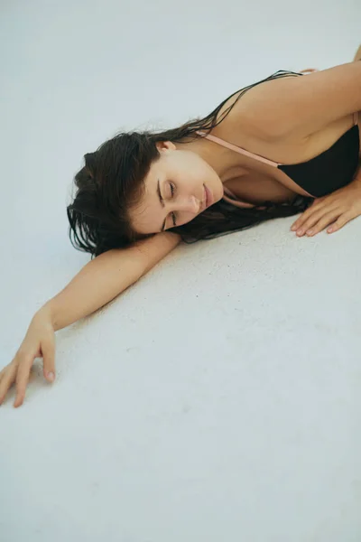 Besada por el sol, mujer morena en bikini negro, modelo sexy con el pelo mojado posando en un resort de lujo, Miami, Florida, EE.UU., fondo borroso, acostado en la superficie blanca, relajación junto a la piscina - foto de stock