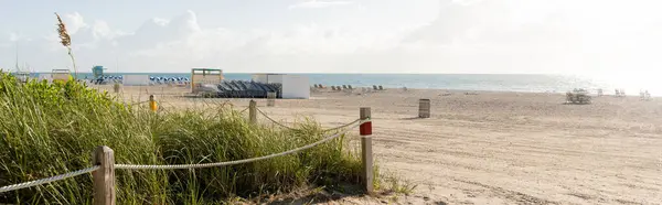 Una spiaggia di sabbia serena fiancheggiata da una recinzione e erba alta, sotto un cielo luminoso, che offre una fuga tranquilla nella natura. — Foto stock