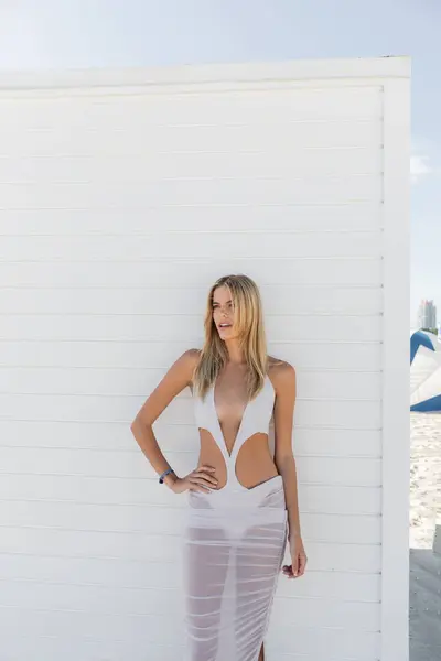 Una joven rubia con un vestido blanco que fluye posa graciosamente al lado de una pared blanca en un entorno de playa en Miami. - foto de stock