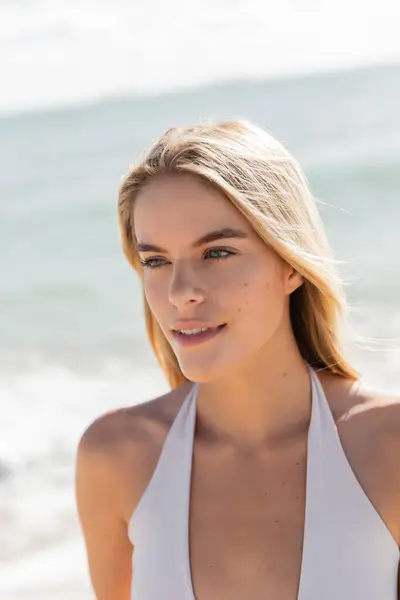 Eine junge, schöne blonde Frau im weißen Bikini steht anmutig am Strand von Miami und umarmt die ruhige Landschaft. — Stockfoto