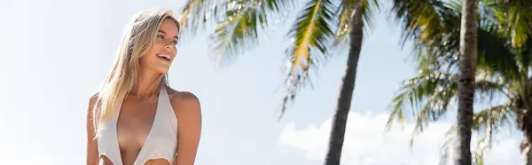 Una mujer rubia impresionante en un bikini blanco se levanta con gracia junto a una palmera alta en una playa de Miami arenosa. - foto de stock