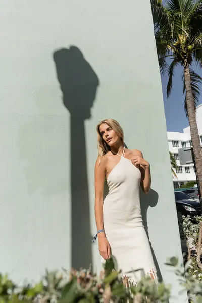 Une superbe jeune femme blonde en robe blanche se tient gracieusement à côté d'un palmier vibrant à Miami. — Photo de stock