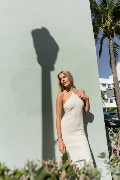 A stunning young blonde woman in a white dress stands gracefully next to a vibrant palm tree in Miami. — Stock Photo