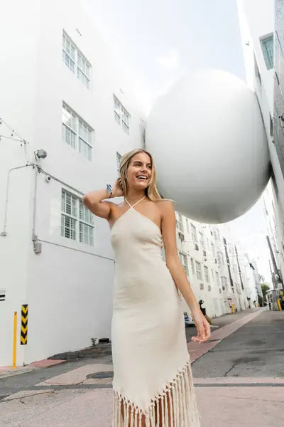 Una joven rubia en un vestido blanco que fluye cerca de una gran bola blanca, exudando belleza y gracia. - foto de stock