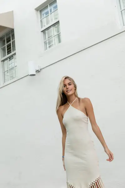 Una joven rubia en un vestido blanco que fluye posa elegantemente frente a un magnífico edificio en Miami. - foto de stock