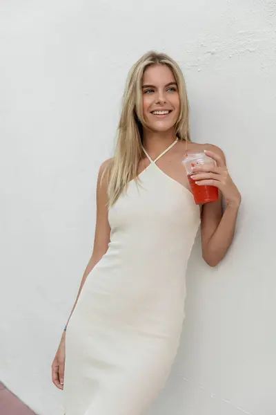 Una joven, hermosa mujer rubia en un vestido blanco que fluye elegantemente sostiene una bebida en un entorno tropical. - foto de stock