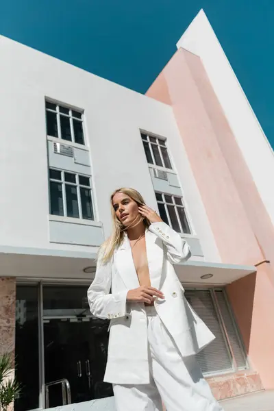 Una mujer rubia impresionante con un traje blanco posando frente a un gran edificio en Miami. - foto de stock