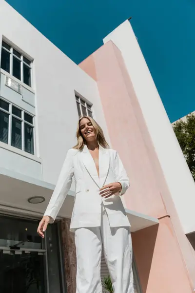Una joven y hermosa mujer rubia se para con confianza en un traje blanco frente a un impresionante edificio de Miami. - foto de stock