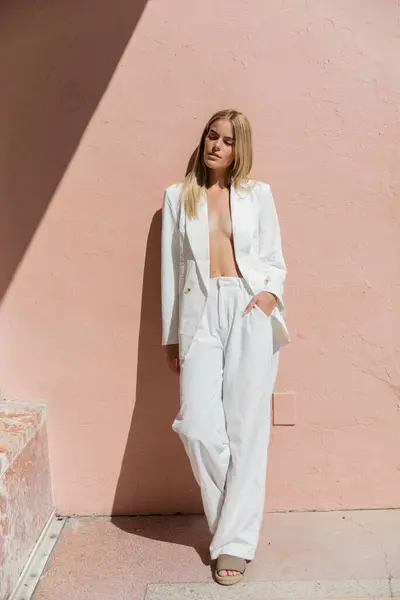 A young, beautiful blonde woman in a white suit gracefully leans against a vibrant pink wall in Miami. — Stock Photo