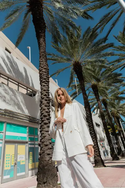 A young, blonde woman stands next to a palm tree on a sunny Miami sidewalk. — Stock Photo