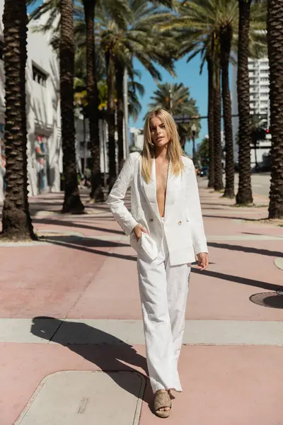 Una mujer rubia exuda confianza mientras camina por una calle de Miami en un impresionante traje blanco, una visión de elegancia. - foto de stock