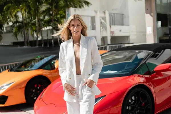 Uma mulher loira deslumbrante fica graciosamente ao lado de um carro esportivo vermelho vibrante em um cenário de Miami glamouroso. — Fotografia de Stock