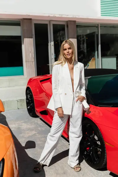 A young, beautiful blonde woman standing confidently next to a sleek red sports car in Miami. — Photo de stock
