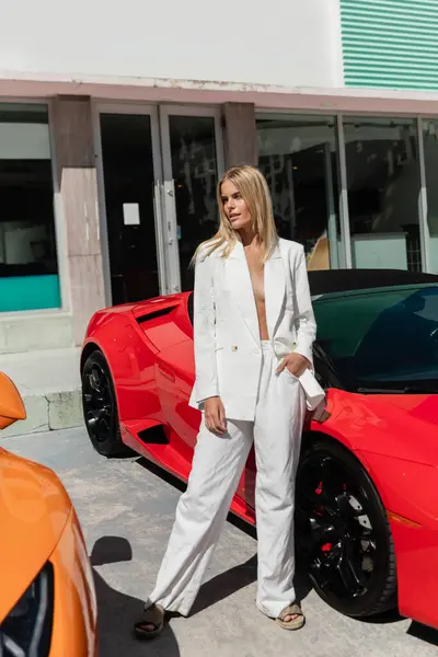 Uma jovem, bela mulher loira de pé confiantemente ao lado de um carro esportivo vermelho vibrante em Miami. — Fotografia de Stock