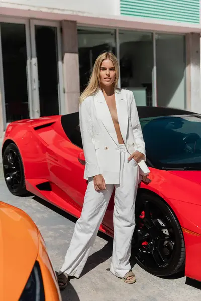 Uma jovem, bela mulher loira fica confiante ao lado de um carro esportivo vermelho vibrante em um cenário ensolarado de Miami. — Fotografia de Stock