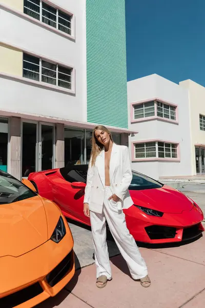 Una giovane, bella donna bionda sta con fiducia accanto a due eleganti auto sportive a Miami. — Foto stock