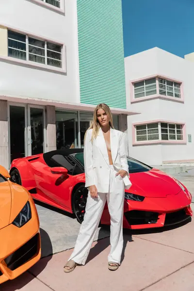 A young blonde woman standing confidently in front of two sleek sports cars. - foto de stock