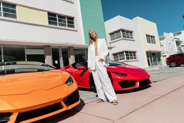 A young blonde woman stands confidently between two cars in front of a building. — Stock Photo