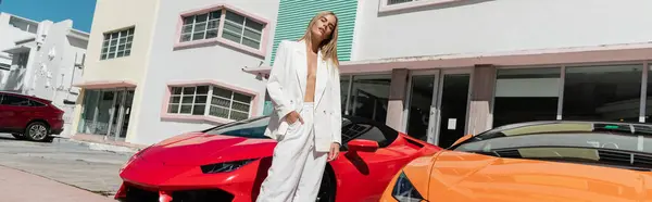 Uma jovem mulher deslumbrante com cabelo loiro de pé elegantemente ao lado de um carro esportivo vermelho vibrante em um cenário de Miami. — Fotografia de Stock