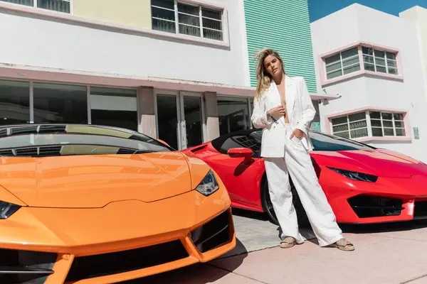 A young blonde woman standing confidently next to two sleek sports cars in Miami. — Stock Photo