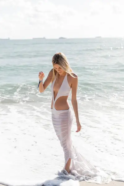 Uma jovem mulher loira está graciosamente em uma praia de areia, olhando para o vasto oceano em Miami.. — Fotografia de Stock